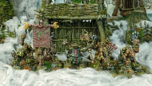 A group of intricately detailed miniatures inspired by Celtic mythology is displayed in front of a rustic, weathered structure covered in moss and foliage. The miniatures, depicting various fantastical creatures, include a central figure holding a banner, flanked by others wielding weapons and adorned with unique features like tusks and intricate patterns. The scene is set amidst a misty, ethereal atmosphere, enhancing the mystical theme of the Famhairean campaign.