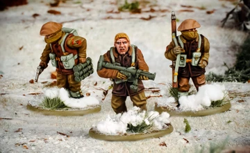 Three multi-part British and Canadian winter infantry figures are depicted in a snowy landscape. The central figure, with short blond hair, is holding a rifle and wearing a brown coat, while the other two figures are dressed in winter gear, one carrying a set of explosives and the other holding a sidearm. The bases of the figures are adorned with patches of snow and tufts of grass, enhancing the wintery scene.
