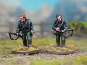 Two detailed miniatures of soldiers from the Sharpe’s Rifles series are displayed on textured bases, set against a blurred natural background. Both figures wear green uniforms with black crossbelts; one holds a rifle and has a serious expression, while the other appears to be in a more contemplative pose. The bases feature tufts of grass, enhancing the realistic terrain effect.