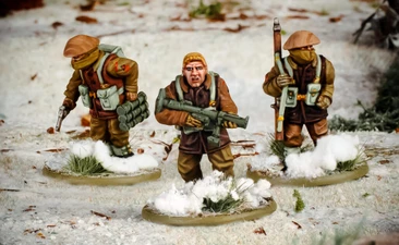 Three multi-part British and Canadian winter infantry figures are depicted in a snowy landscape. The central figure, with short blond hair, is holding a rifle and wearing a brown coat, while the other two figures are dressed in winter gear, one carrying a set of explosives and the other holding a sidearm. The bases of the figures are adorned with patches of snow and tufts of grass, enhancing the wintery scene.