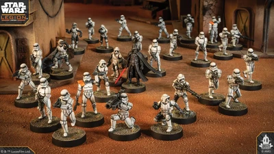 A group of Star Wars miniatures features several Stormtroopers in white armor, positioned in a desert-like environment with a backdrop of a weathered building. Among them stands a prominent figure in black armor, holding a red lightsaber, likely representing Darth Vader. The scene is detailed with various poses of the troopers, some aiming weapons, while others appear to be in action, all set on circular bases with textured surfaces.
