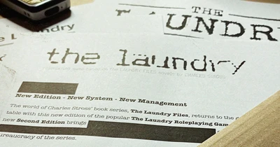 A close-up view of a printed document featuring the title "THE LAUNDRY" prominently displayed at the top, with a distressed font style. Below the title, the text indicates that it is based on the "LAUNDRY FILES" novels by Charles Stross, and mentions "System - New Management." The document appears to be part of promotional material for the latest edition of the Laundry RPG.