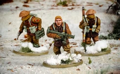 Three multi-part British and Canadian winter infantry figures are depicted in a snowy landscape. The central figure, with short blond hair, is holding a rifle and wearing a brown coat, while the other two figures are dressed in winter gear, one carrying a set of explosives and the other holding a sidearm. The bases of the figures are adorned with patches of snow and tufts of grass, enhancing the wintery scene.