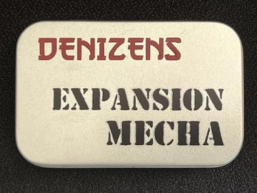 A small, silver tin features the text "DENIZENS" in bold red letters at the top and "EXPANSION MECHA" in large, black, stencil-style font below. The tin's surface has a smooth, metallic finish, and it is set against a dark background, emphasizing its compact and portable design. This packaging represents the new expansion for the Space Denizens wargame, which includes mechs and terrain features.
