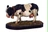 A detailed miniature of a black and white cow stands on a textured base resembling grass and dirt. The cow is depicted with a collar and bell around its neck, showcasing realistic features such as its distinct markings and posture. This figure adds a whimsical touch, fitting for a fantasy tavern setting.