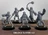 A group of five Drudge Slayer miniatures stands on circular bases, each showcasing unique designs and weaponry. The figures are muscular and armored, featuring various mechanical elements and menacing expressions. They are posed in dynamic stances, with some wielding large weapons like spiked flails and others displaying intricate details in their armor, all rendered in a gray color that highlights their features against a simple background.
