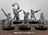 A collection of five Drudge Slayer miniatures stands on a wooden surface, each intricately designed with a mix of armor and weaponry. The figures feature various poses, showcasing unique details such as spiked weapons, chains, and mechanical elements, all rendered in a gray color. The text "DRUDGE SLAYERS F-J" is displayed prominently at the bottom of the image.