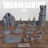 A collection of 3D printed terrain pieces from the Warmachine "Old Umbrey Terrain Set" is displayed on a wooden surface. The set includes a large, intricately designed building with pointed towers, a tall stone tower, and several smaller terrain elements like rocky outcrops, trees, and fences. Various bases and scatter terrain pieces are arranged around the central structures, showcasing a mix of natural and architectural features, all in a gray color. A pair of dice is also visible in the foreground.