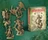 A collection of intricately designed miniatures from the Dragonbane RPG is displayed against a textured green background. The set includes a variety of characters, such as a knight with a spear, a dragon, and a sorcerer, all rendered in a beige color, showcasing their detailed features. To the right, a box labeled "DRAGONBANE DEMONS SET" features artwork of a menacing creature, indicating the theme of the miniatures within the set.