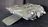 A large, detailed model of a spaceship, featuring a flat, ramp-like structure at the front and a complex array of panels and textures across its surface. The ship has a robust, angular design with various protrusions and openings, suggesting functionality for cargo or troop deployment. The model is presented in a light gray color against a dark background, highlighting its intricate details.