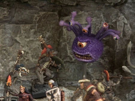 A collection of fantasy miniatures features several lizardmen, one wielding a shield and weapon, and another with a torch, set against a rocky backdrop. In the background, a large, purple creature with multiple eyes and a menacing expression looms, adding to the scene's fantastical atmosphere. A female character in dark robes stands nearby, while other figures in armor are partially visible, enhancing the sense of adventure.