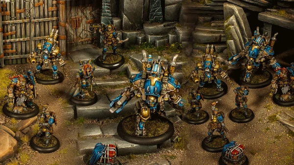 A group of Cygnar miniatures is depicted in a detailed tabletop setting, showcasing the Gravedigger Auxiliaries unit. In the foreground, a character heavy warjack named Beaker stands prominently, adorned in blue and gold armor, with various mechanical features. Surrounding it are several elite veterans, including a female commander with red hair and a rifle, an ogrun captain, and a unit of Gravedigger Commandos, all featuring a mix of blue and gold uniforms, set against a backdrop of stone steps and a weathered door.