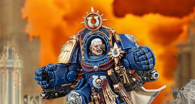 A detailed miniature of Marneus Calgar, a prominent character from the Warhammer 40,000 universe, is depicted in a striking pose. He wears ornate blue power armor adorned with gold embellishments, featuring a prominent Ultramarines symbol on his chest and a skull motif. The background is a dramatic orange, suggesting a fiery battlefield, enhancing the intensity of the scene.
