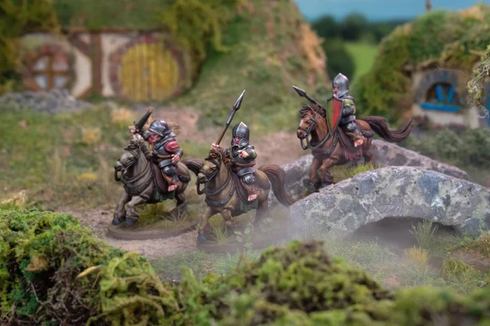 Three mounted halfling cavalry figures are depicted, each riding stout ponies and equipped with spears. The riders wear armor and helmets, showcasing a mix of colors, including silver and red accents. The scene is set in a lush, grassy landscape with rocky terrain and a distant background of rolling hills, creating a vibrant and dynamic atmosphere.