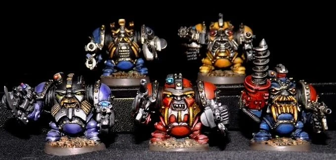Five multipart, white metal miniatures of armoured dwarves are displayed against a dark background. The front row features two dwarves in vibrant blue and red power armour, each holding different weapons, while the back row showcases three more dwarves in a mix of gold and metallic colours, also armed. Each miniature is intricately detailed, with various mechanical elements and unique designs, highlighting the sci-fi and Victoriana influences.