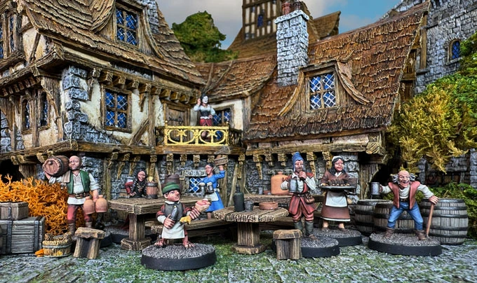 A detailed scene depicts a bustling tavern setting with several 28mm scale white metal miniatures representing various townsfolk. Characters include a barmaid serving drinks, a bard playing music, and patrons engaged in conversation, all set against a backdrop of a rustic tavern with a thatched roof and stone walls. The environment features wooden tables, barrels, and vibrant foliage, creating a lively atmosphere typical of a fantasy RPG setting.