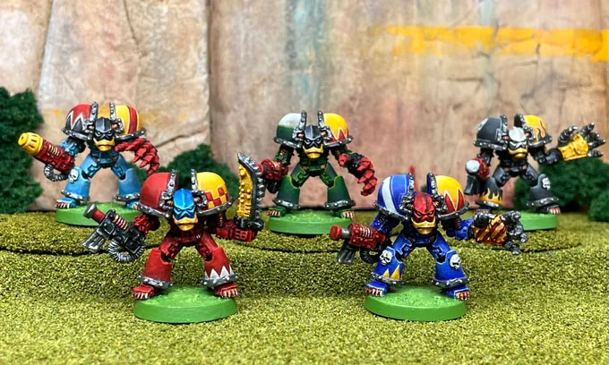 A group of five colorful, 28mm scale space orc miniatures stands on a grassy terrain, each equipped with various weapons and wearing distinctive armor. The orcs are painted in vibrant colors, including red, blue, and green, with unique designs on their helmets and armor, showcasing their cybernetic enhancements. They are posed dynamically, suggesting readiness for battle, against a backdrop of rocky terrain and greenery.