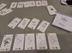 A tabletop game setup featuring a collection of beautifully illustrated cards from the game "Florilegium." The cards display various botanical illustrations, including flowers and plants, with delicate details and soft colors. Some cards are arranged in a grid pattern, while others are stacked, alongside a box labeled "Florilegium," all set on a neutral-colored table.