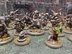 A group of anthropomorphic animal miniatures, primarily featuring badger-like figures, is depicted in a dynamic scene. The miniatures are intricately painted, showcasing a variety of colors and details, with some wearing armor and wielding weapons, while others appear to be casting spells or holding shields. The background features a textured terrain with grass and rocks, enhancing the fantasy setting, and a stone wall is visible in the distance, adding depth to the scene.