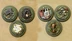 Four objective markers are displayed on a textured surface. Each marker features a distinct design: one shows a collection of tools, another depicts a hand emerging from a burrow, a third features a map with a castle and trees, and the last displays a shield with a checkered pattern. The markers are set against a green, grassy background, enhancing their thematic connection to the Burrows and Badgers game.