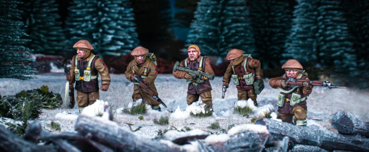 Three British and Canadian winter infantry figures are depicted in a snowy landscape, surrounded by patches of grass and rocks. Each figure is dressed in warm winter gear, with various poses: one holds a rifle, another has a sidearm, and the third is equipped with a machine gun. The background features dark green trees, enhancing the wintery atmosphere of the scene.