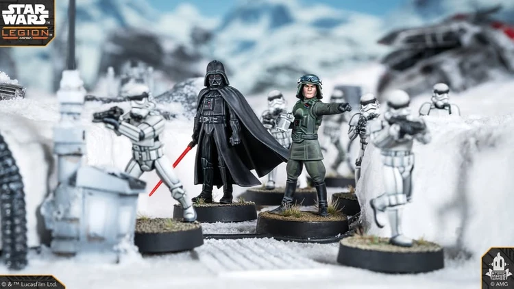 A detailed scene from Star Wars Legion features iconic miniatures set in a snowy landscape. Prominently displayed are Darth Vader, holding a red lightsaber, and a uniformed officer pointing, surrounded by several Stormtroopers. The terrain includes patches of grass and rocky elements, enhancing the immersive atmosphere of the battlefield.