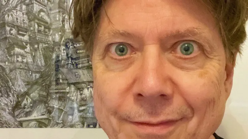 A close-up portrait of a man with light brown hair and striking green eyes, smiling slightly. He is wearing a black shirt and is positioned in front of a detailed, artistic background featuring a complex, fantastical structure, possibly a building or landscape. The overall tone of the image is casual and inviting, with a focus on the subject's expressive features.