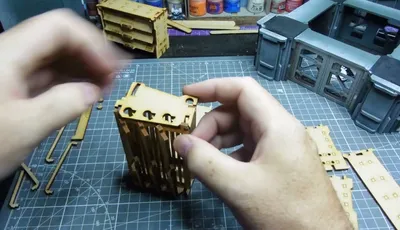 Hands assembling a wooden model on a cutting mat with tools and paint jars in the background.