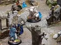 A whimsical scene features a fantastical creature resembling a goat or sheep with large horns, sitting on a rocky terrain adorned with greenery and small flowers. Nearby, a character in a blue cloak holds a sign that reads, "Tendo is cheap and illegal! Not lying!" while another figure stands beside them, holding a sword. Blue dice are scattered throughout the scene, adding to the gaming atmosphere.