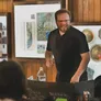 A smiling game master stands behind a black game screen, engaging with players at a tabletop gaming event. He wears a black shirt and glasses, with a water bottle on the table beside him. The background features wooden walls adorned with various artworks and decorative plates, creating a warm, inviting atmosphere for the gaming session.