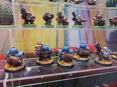 A display of miniature figures showcases a lineup of small, resin models resembling futuristic space dwarves, known as Roknauts. The front row features five figures in blue armor, each equipped with various weapons, standing on textured bases with a sandy appearance. Behind them, additional miniatures are visible, depicting a variety of characters and creatures, set against a colorful backdrop.