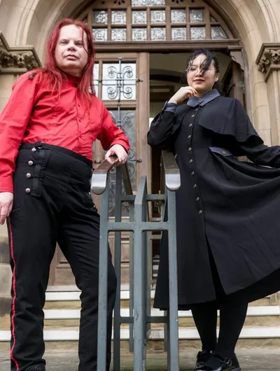Two individuals pose in front of a grand, gothic-style building. One person, wearing a bright red shirt and black trousers with red stripes, stands confidently with arms crossed. The other person, dressed in a long black coat with a high collar and a blue shirt underneath, holds the hem of their coat, creating a dramatic silhouette. The ornate entrance of the building, featuring large wooden doors and intricate stonework, serves as a striking backdrop.