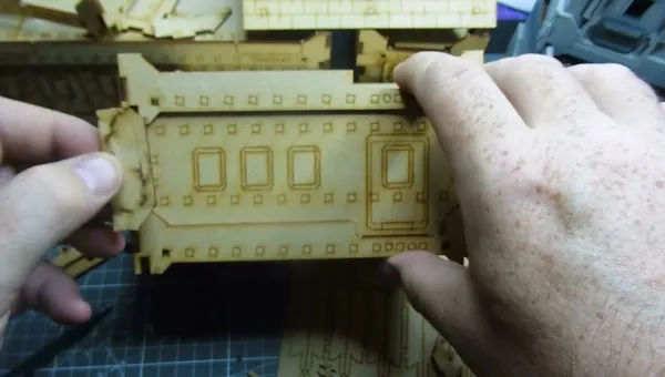 A hand holds a light green plastic model building piece, designed to resemble a fortified or industrial structure. The piece features four rectangular window cutouts arranged in two rows, each window framed with raised borders. Along the top edge, small circular holes are evenly spaced. The model appears to be part of a modular terrain set, resting on a patterned surface. The background shows additional similar model pieces, slightly out of focus. No text is visible on the model.