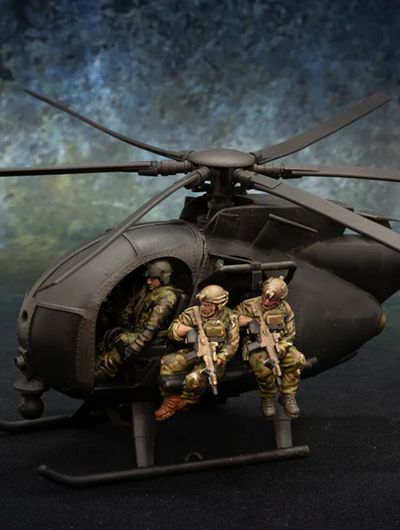 A detailed model of a Little Bird helicopter, featuring a dark color scheme, is prominently displayed. Two miniature soldiers are seated on the helicopter's skids, equipped with rifles, while another soldier is visible inside the cockpit. The background features a textured gradient, enhancing the focus on the helicopter and its crew.