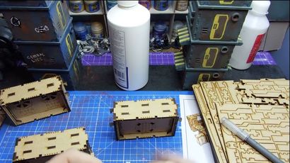 A workbench scene features a partially assembled laser-cut building structure made of light wood, positioned in the foreground. Behind it, a stack of completed structures in various colors, including blue and yellow, is visible, alongside bottles of paint and glue. A cutting tool rests on the table, and a printed reference image of a similar structure is partially visible.