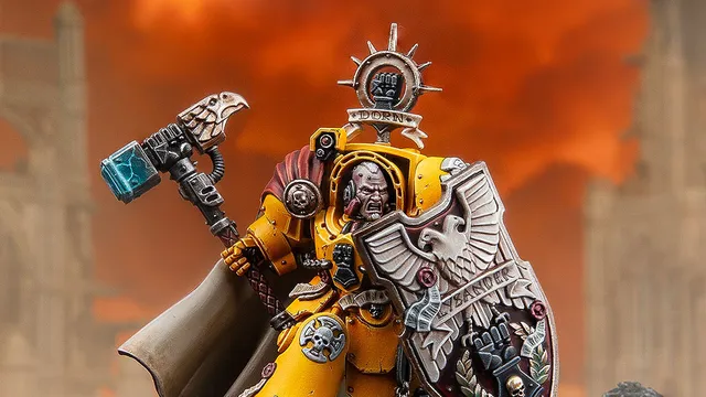 A dynamic miniature of Darnath Lysander in bright yellow Terminator armour, holding the relic storm shield Rampart in one hand and the Fist of Dorn power hammer in the other. His expression is fierce, and he wears a flowing cape, with intricate details on his armour and shield, including the name "LYSANDER" and various symbols. The background features a dramatic, fiery sky, enhancing the imposing presence of the character.