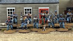 A group of ten detailed 28mm scale miniatures representing dismounted US Cavalry and Buffalo Soldiers stands in front of a wooden building with a red door. The miniatures are posed with various weapons, including rifles and a bugle, showcasing a mix of uniforms and accessories, such as scarves and hats. A small beagle and a cactus are also present among the figures, all mounted on 25mm round bases with a sandy terrain effect.