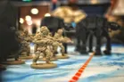 A group of miniature soldiers in beige uniforms, armed and poised for action, are positioned in the foreground on a tabletop battlefield. In the background, two large, black AT-AT walkers loom, creating a stark contrast against the lighter colors of the soldiers. The scene is set on a blue terrain mat, with a red line marking a path across the battlefield.