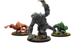 The image features three miniatures from Modiphius’ Fallout: Wasteland Warfare line. On the left is a snallygaster, a hexapod creature with a reddish-brown body, multiple eyes on its back, and a long, protruding tongue. In the center stands a large, muscular megasloth, covered in gray fur, with a menacing pose and a raised arm. To the right is another creature, possibly a variant of the snallygaster, in green, with a textured body and large eyes, all set on circular bases adorned with tufts of grass.