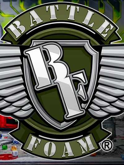 Stylized emblem featuring a shield design. At the top of the shield, bold white capital letters spell "BATTLE". In the center, a large white number "3" is integrated with a sword pointing downward. Below the shield, a green banner with white capital letters reads "FOAM" and to the right of it, a registered trademark symbol "(R)". The background depicts a cluttered tabletop with miniature terrain pieces, small figures, dice, and a red vehicle model. The setting appears to be a gaming or hobby workspace.