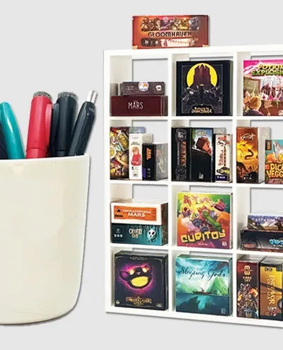 A white ceramic cup filled with pens and a stapler sits on the left. To the right is a white grid-style shelf holding multiple small board game boxes, including visible titles such as "GLOOMHAVEN", "MARS", "POTION EXPLOSION", "DICED VEGGIES", "TERRAFORMING MARS", "CUBITOY", "Sleeping Gods", and others.