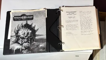 A three-ring binder containing the playtest manuscript for "ManaStorm: The Magic Adventure Game," featuring a black and white cover illustration of a character with a spiked helmet and a weapon. The title "MANASTORM The Magic Adventure Game" is prominently displayed at the top, along with the version number and date on the first page inside the binder. Additional pages include loose character sheets and player handouts, indicating the game's mechanics and spells.