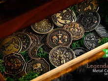 A wooden box filled with metal coins featuring intricate designs, including symbols like a crescent moon, crowns, and shields. Green moss-like material lines the box. Overlay text reads: "FUNDED IN 2 HOURS" and "Legendary Metal Coins Season 9."