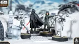 A detailed scene features Darth Vader standing prominently with his red lightsaber drawn, dressed in his iconic black armor and cape. Flanking him are two Stormtroopers, one aiming a blaster, set against a snowy landscape with scattered debris and structures. The miniatures are mounted on circular bases, showcasing their intricate designs and the dynamic pose of the characters.