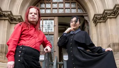 Two individuals pose in front of a grand, gothic-style building. One person, wearing a bright red shirt and black trousers with red stripes, stands confidently with arms crossed. The other person, dressed in a long black coat with a high collar and a blue shirt underneath, holds the hem of their coat, creating a dramatic silhouette. The ornate entrance of the building, featuring large wooden doors and intricate stonework, serves as a striking backdrop.