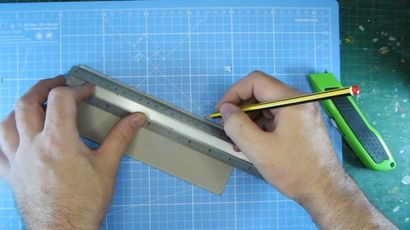 A hand is holding a yellow pencil against a metal ruler, positioned on a piece of cardboard on a cutting mat. The cutting mat features a grid pattern with measurements in centimeters, and a green utility knife is visible nearby. The scene suggests preparation for cutting or crafting a new paint shelf.