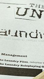 A close-up view of a printed document featuring the title "THE LAUNDRY" prominently displayed at the top, with a distressed font style. Below the title, the text indicates that it is based on the "LAUNDRY FILES" novels by Charles Stross, and mentions "System - New Management." The document appears to be part of promotional material for the latest edition of the Laundry RPG.