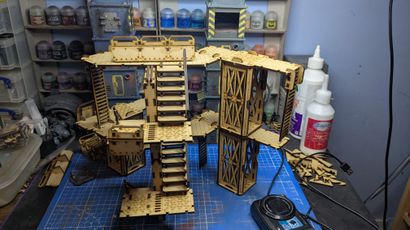 A detailed assembly of modular terrain pieces made from laser-cut wood, featuring multiple levels, staircases, and platforms. The structure includes various textures and patterns, with a mix of open and enclosed areas, showcasing a futuristic industrial design. In the background, paint bottles and tools are visible, indicating a workspace for painting and customizing the terrain.