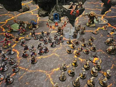 Miniature dark fantasy wargame scene with two armies on a cracked, lava-like battlefield. Left side features dwarven figures with armor and weapons, including a large blue-robed figure with a glowing orb.