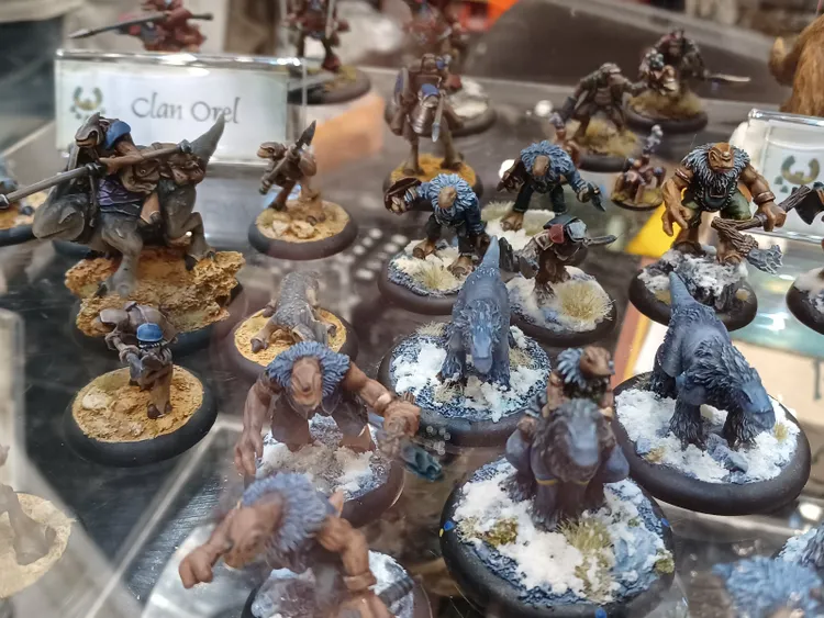 A collection of fantasy miniatures displayed in a glass case, featuring various anthropomorphic figures and creatures. Prominent are several bear-like warriors with blue fur and weapons, alongside smaller creatures resembling wolves and lizard-like beings. The bases of the miniatures are textured with snow and grass, enhancing the thematic presentation of a wintery landscape.